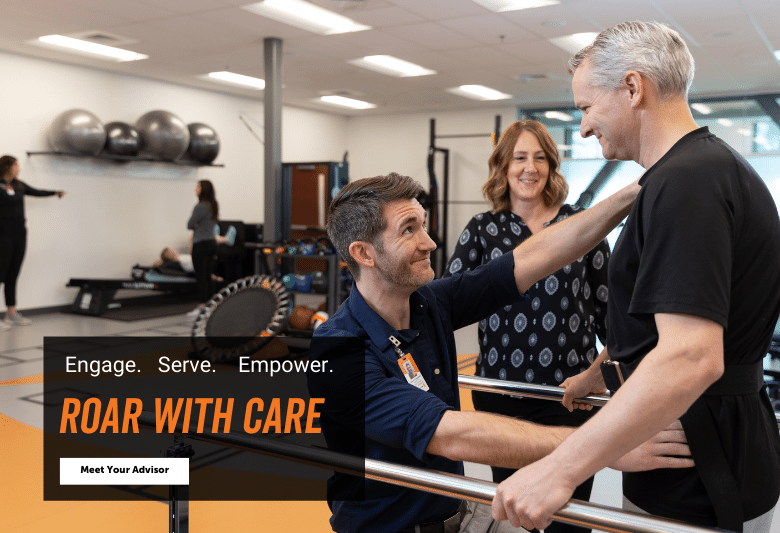 A physical therapist in a navy blue polo holds onto an older man to assist him in movement. The older man is standing and holding onto bars on either side of him. A woman is watching and smiling in the background. Text box reads: Engage. Serve. Empower. Roar with care. Button reads: Meet your advisor.